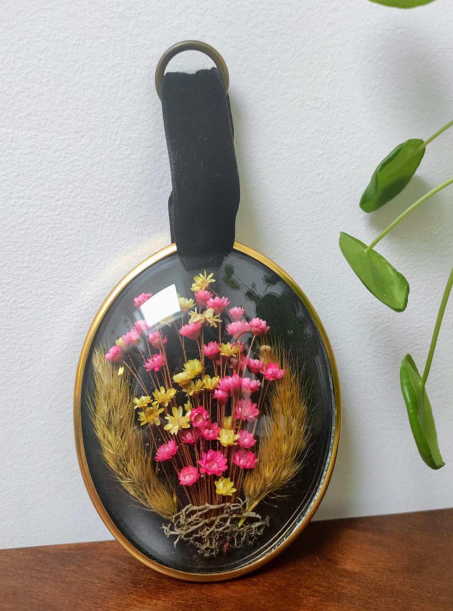 Beau petit cadre vintage de forme ovale avec fleurs naturelles séchées en bouquet. Le verre bombé est bordé d’un liseré doré. Une attache en velours est présente. De belles couleurs et beaucoup de charme pour ce beau petit cadre.