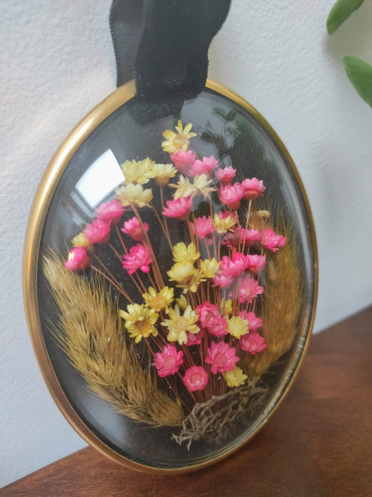 Beau petit cadre vintage de forme ovale avec fleurs naturelles séchées en bouquet. Le verre bombé est bordé d’un liseré doré. Une attache en velours est présente. De belles couleurs et beaucoup de charme pour ce beau petit cadre.