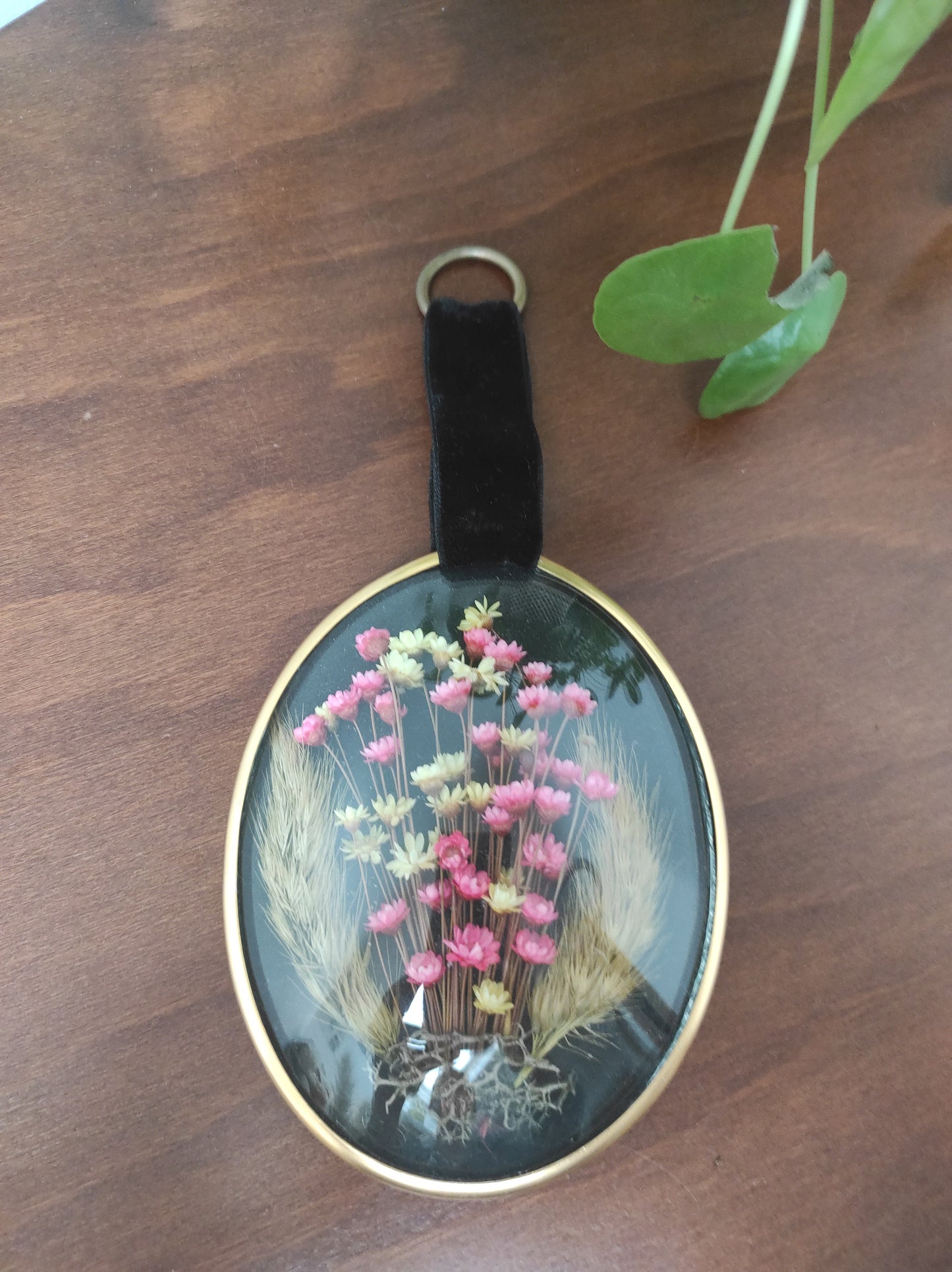 Beau petit cadre vintage de forme ovale avec fleurs naturelles séchées en bouquet. Le verre bombé est bordé d’un liseré doré. Une attache en velours est présente. De belles couleurs et beaucoup de charme pour ce beau petit cadre.