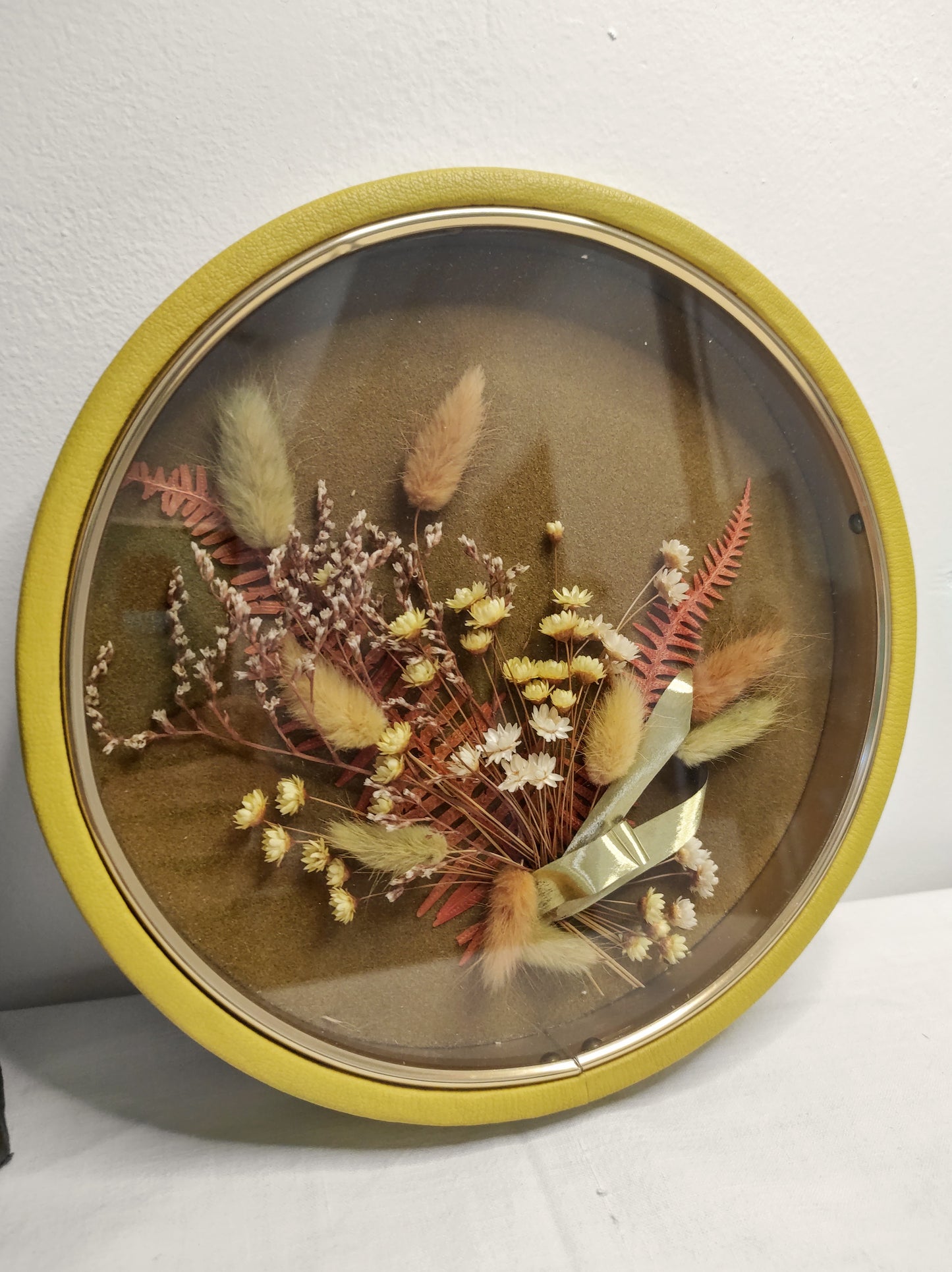 Cadre de fleurs naturelles séchées vintage. Beau Cadre vintage de forme ronde avec fleurs naturelles séchées en bouquet. Le verre est bordé d’un liseré doré. Le contour est de couleur jaune moutarde épais style simili cuir. Une attache est présente au dos. De belles couleurs automnales et beaucoup de charme pour ce beau cadre. Cadre vintage
