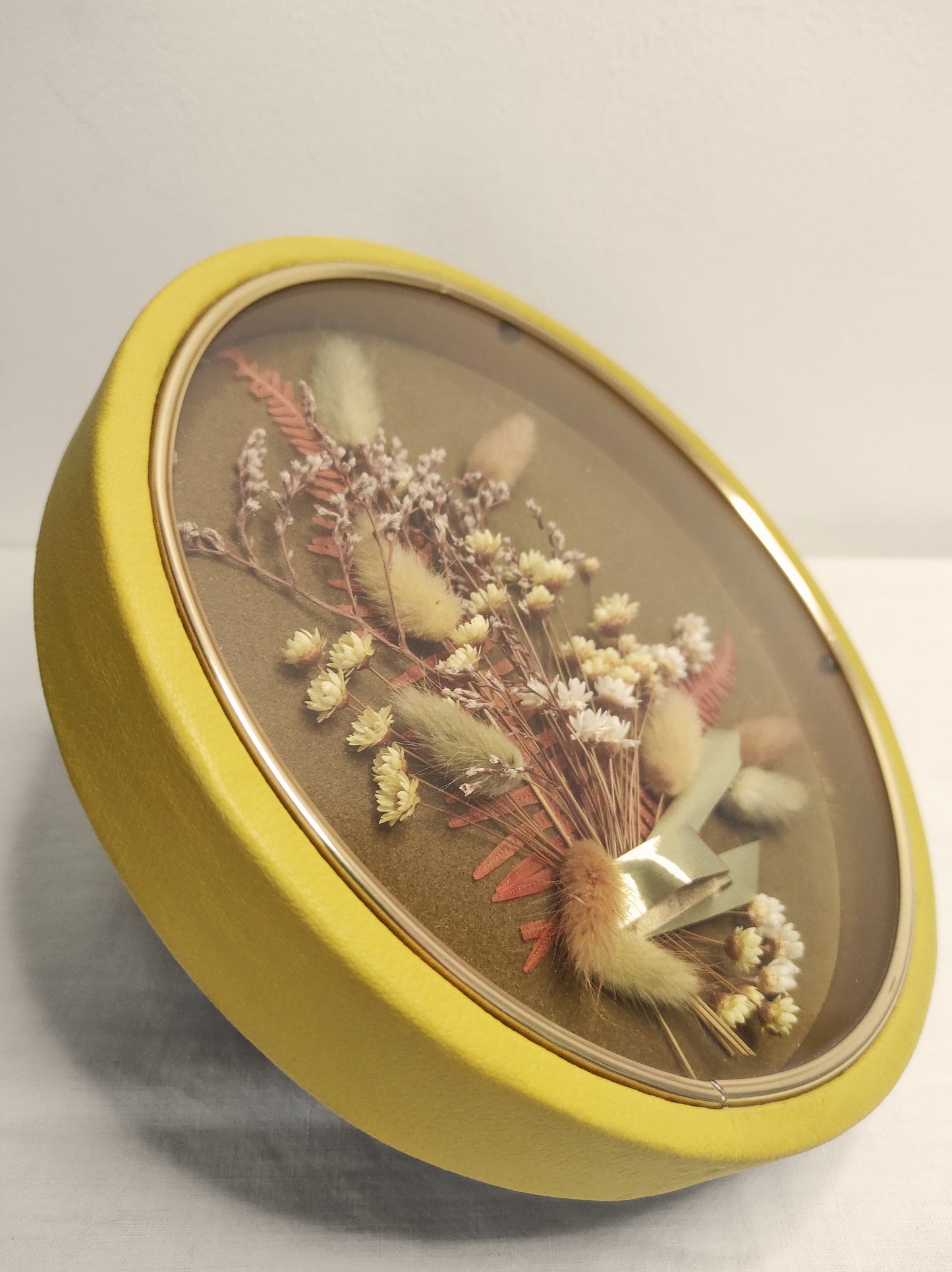 Cadre de fleurs naturelles séchées vintage. Beau Cadre vintage de forme ronde avec fleurs naturelles séchées en bouquet. Le verre est bordé d’un liseré doré. Le contour est de couleur jaune moutarde épais style simili cuir. Une attache est présente au dos. De belles couleurs automnales et beaucoup de charme pour ce beau cadre. Cadre vintage