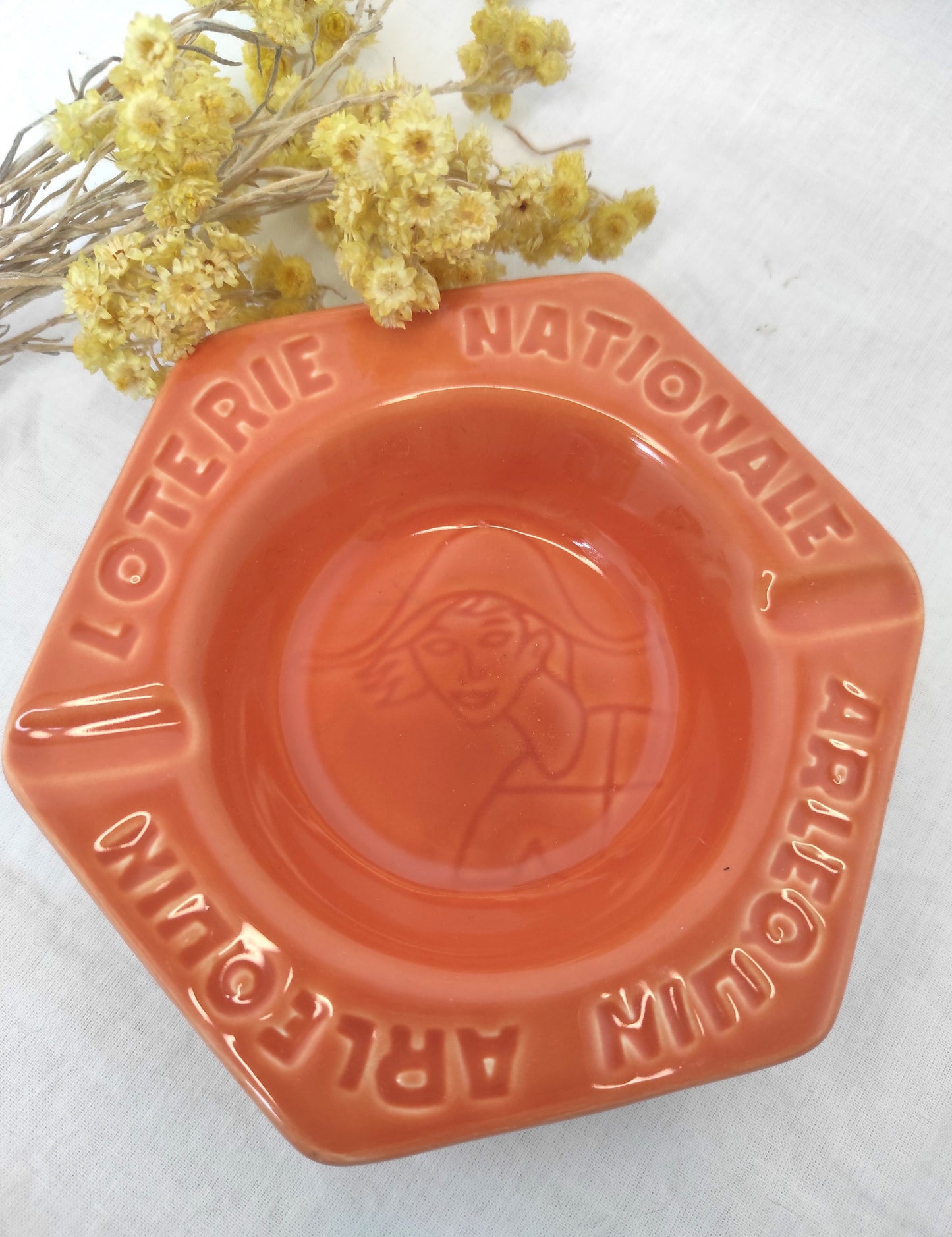 Ancien cendrier publicitaire LOTERIE NATIONALE ARLEQUIN. Joli petit cendrier publicitaire vintage Loterie Nationale Arlequin en faïence de couleur orange saumon en très bon état (aucun éclat). Il fera une superbe déco rétro et colorée sur une étagère ou un joli vide poche sur une commode ! Décoration vintage