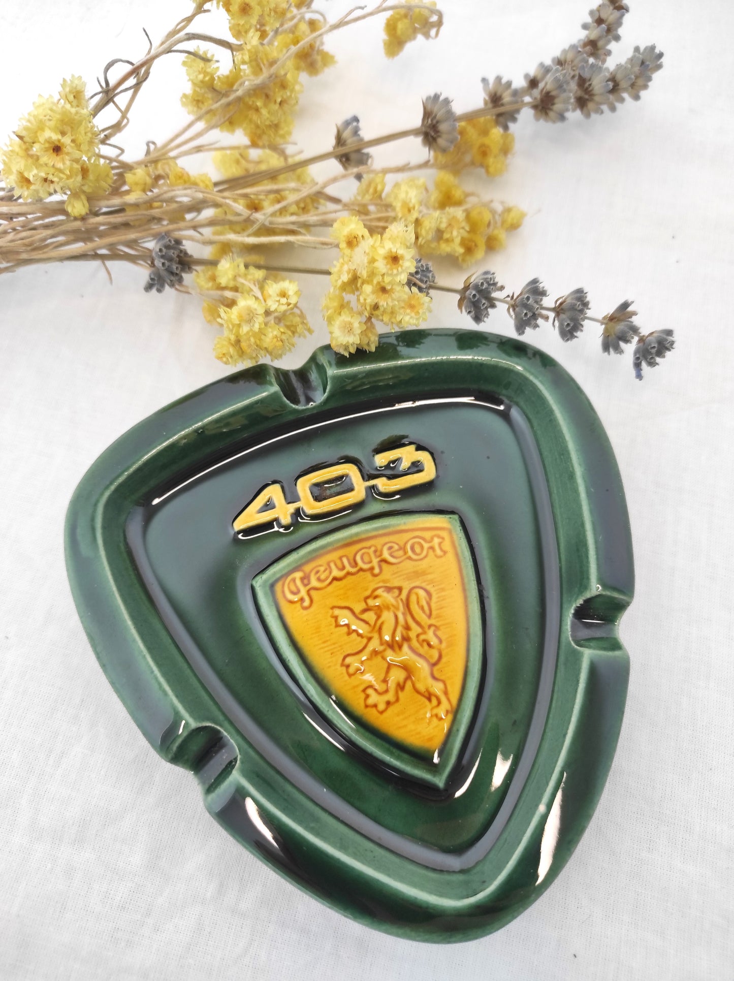 Rare cendrier publicitaire des années 60 PEUGEOT 403. RARE cendrier publicitaire des années 60 pour la Peugeot 403 en faïence de couleur vert sapin et jaune moutarde. Il fera une superbe déco rétro et colorée sur une étagère ou un joli vide poche sur une commode ! A noter : un mini éclat à peine visible (voir photo) mais en très bon état vu son âge, signé au dos. Décoration vintage.
